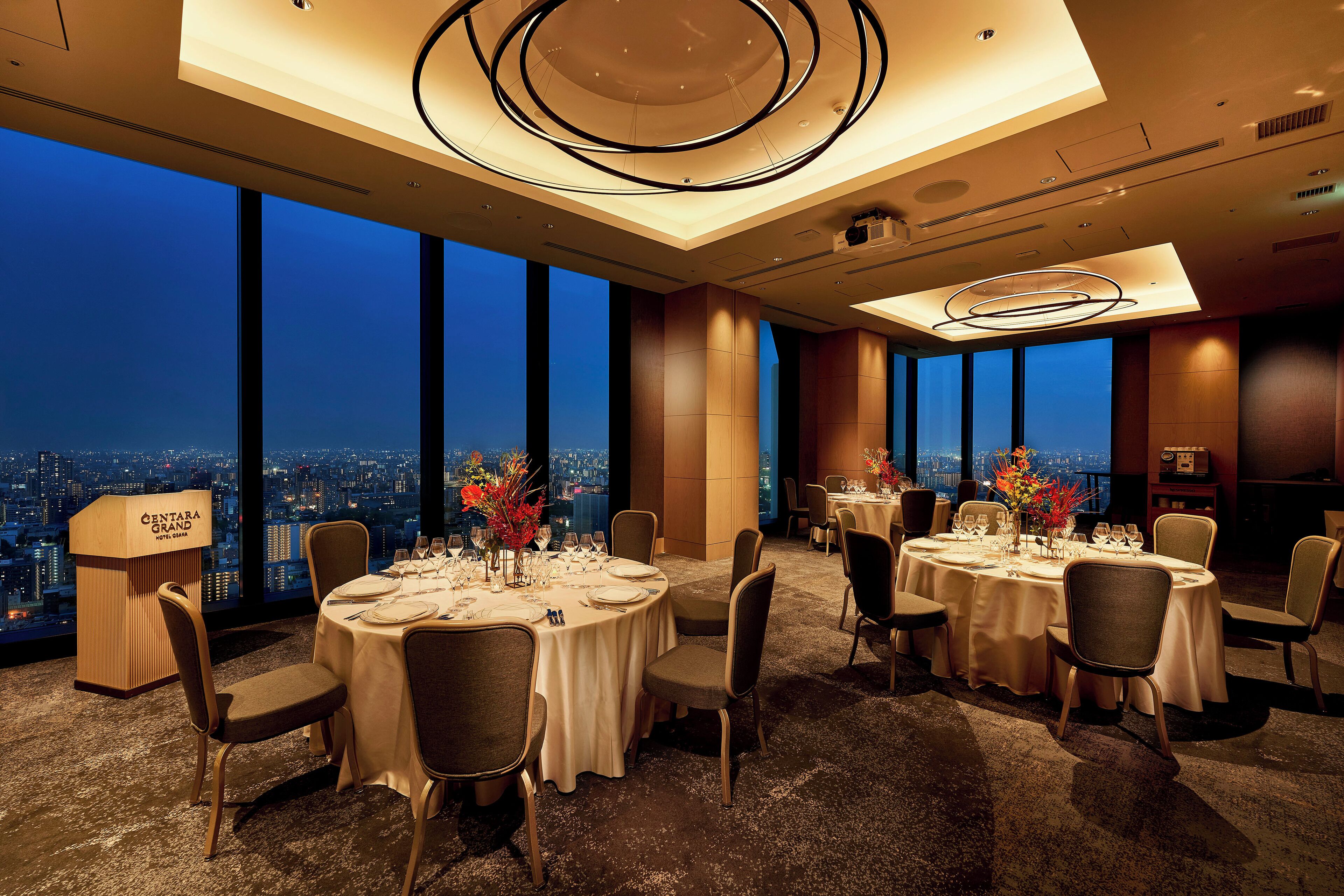 A high-rise hotel dining area with multiple round tables set for dining, large windows offering city views, and a modern ceiling design.