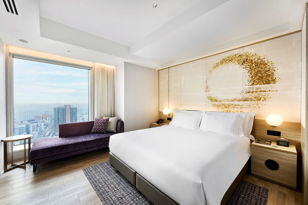A modern hotel room with a large bed, a city view, a sofa, and a nightstand with a lamp.