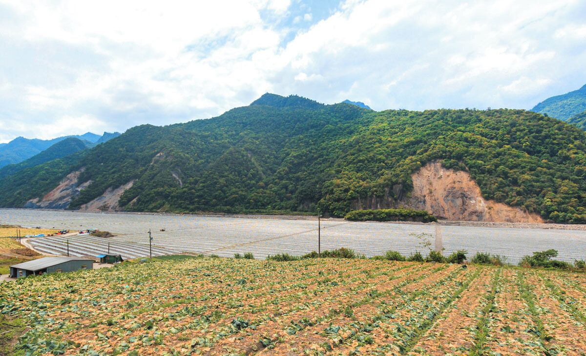 Un río con un puente, un campo de cultivo y una cadena montañosa al fondo.