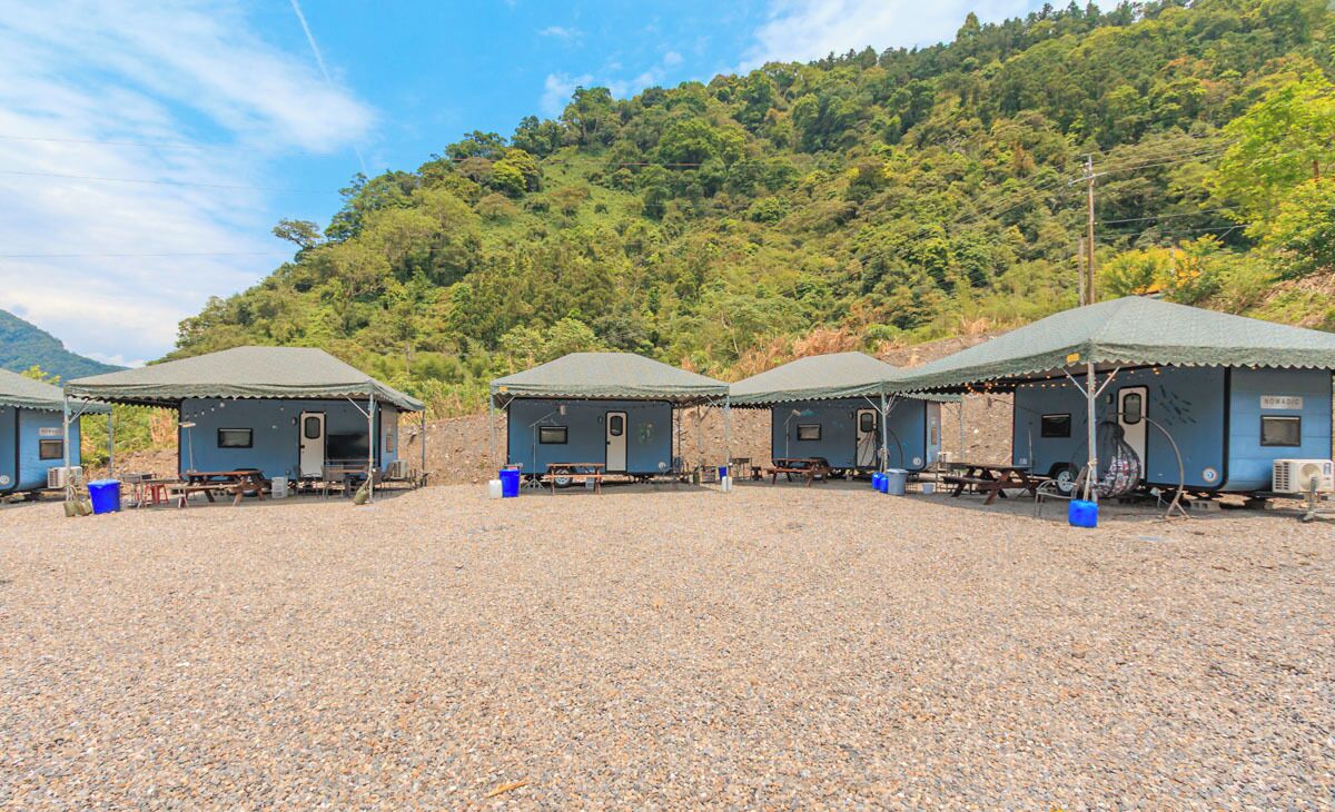 A row of small, blue cabins with metal roofs, arranged in a gravel area with a forested hill in the background.