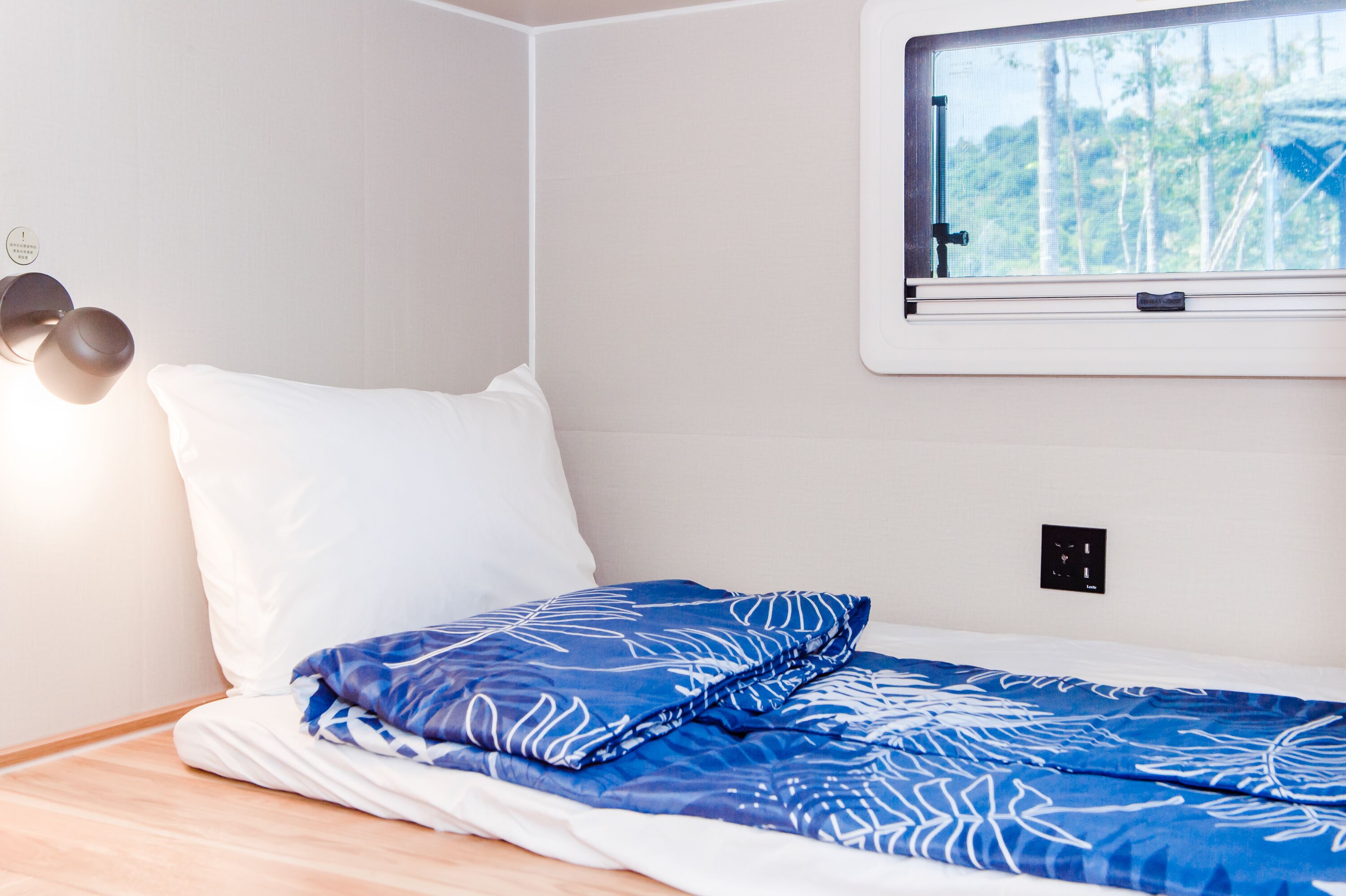 Ein Bett mit weißer und blauer Bettwäsche, ein Fenster mit Blick auf Bäume und eine Wandleuchte.