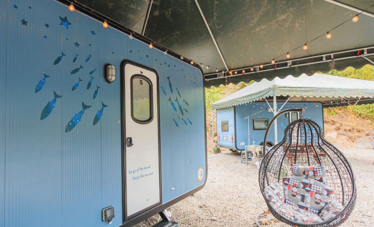 A blue RV with a door labeled "let go of the world. Enjoy the sea and" and a hanging chair with patterned cushions.