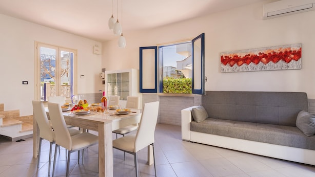 Un salon moderne avec un canapé gris, un coin repas avec une table et des chaises blanches, et une grande fenêtre offrant une vue dégagée.