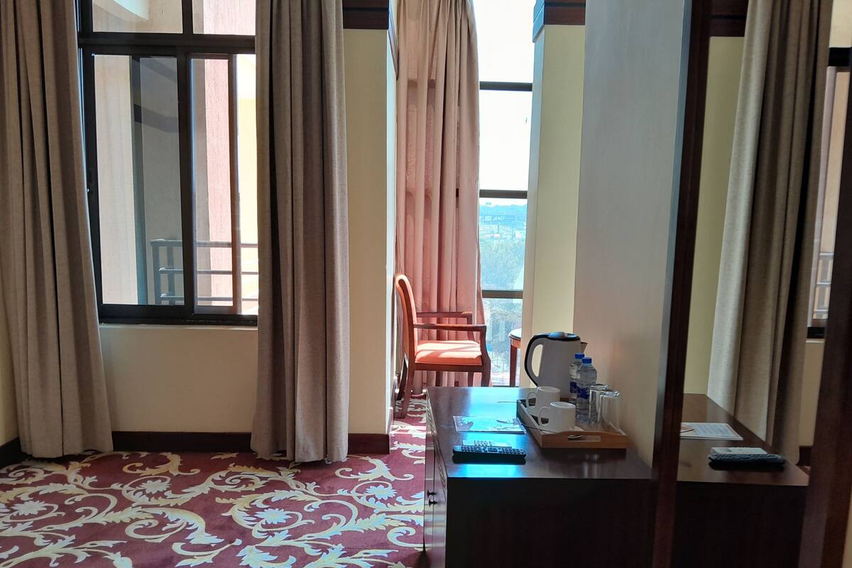 Una habitación con alfombra estampada, un escritorio con teclado, una silla y vistas a un edificio a través de una ventana.