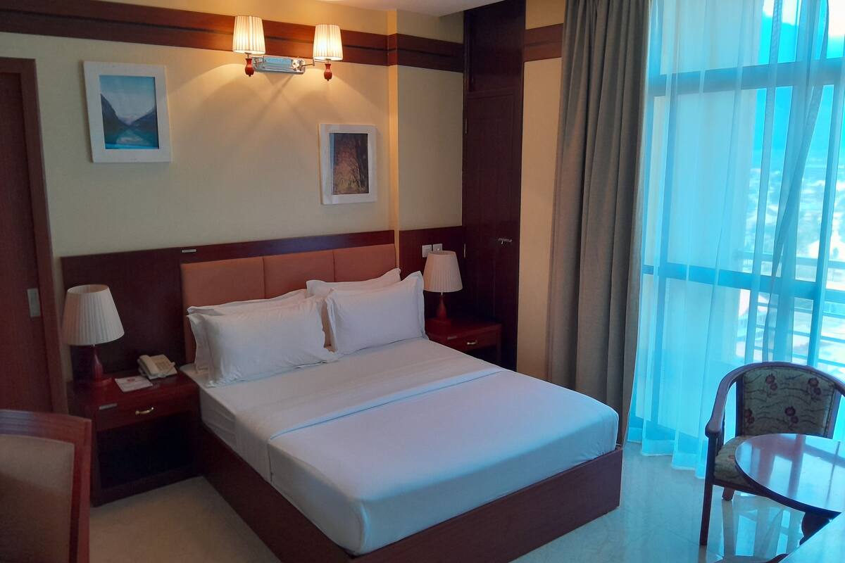 Habitación de hotel con una cama grande, dos mesitas de noche con lámparas, una silla y vistas al exterior a través de cortinas translúcidas.