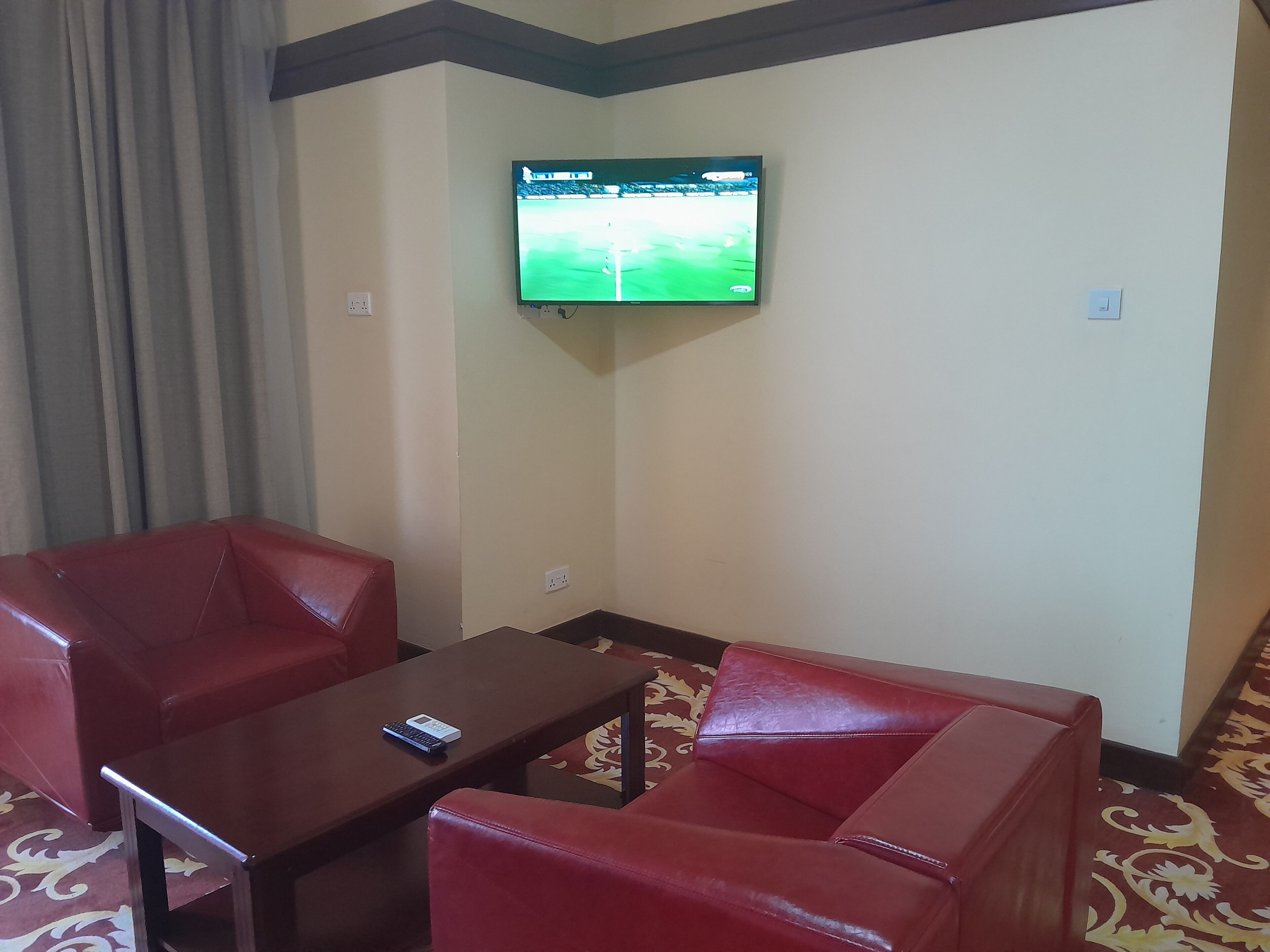 Ein Hotelzimmer mit an der Wand montiertem Fernseher, einem braunen Tisch, zwei roten Ledersesseln und einem gemusterten Teppich.