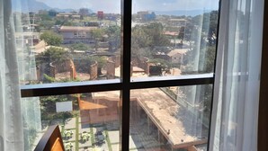 Vista a través de una ventana con cortinas, que muestra una zona residencial con casas, árboles y una cadena montañosa distante.