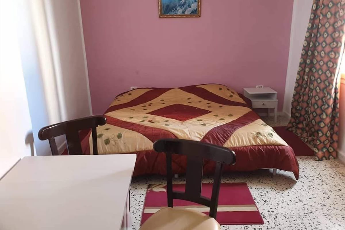 A bedroom with a bed, a chair, a small bedside table, and a painting on the wall.