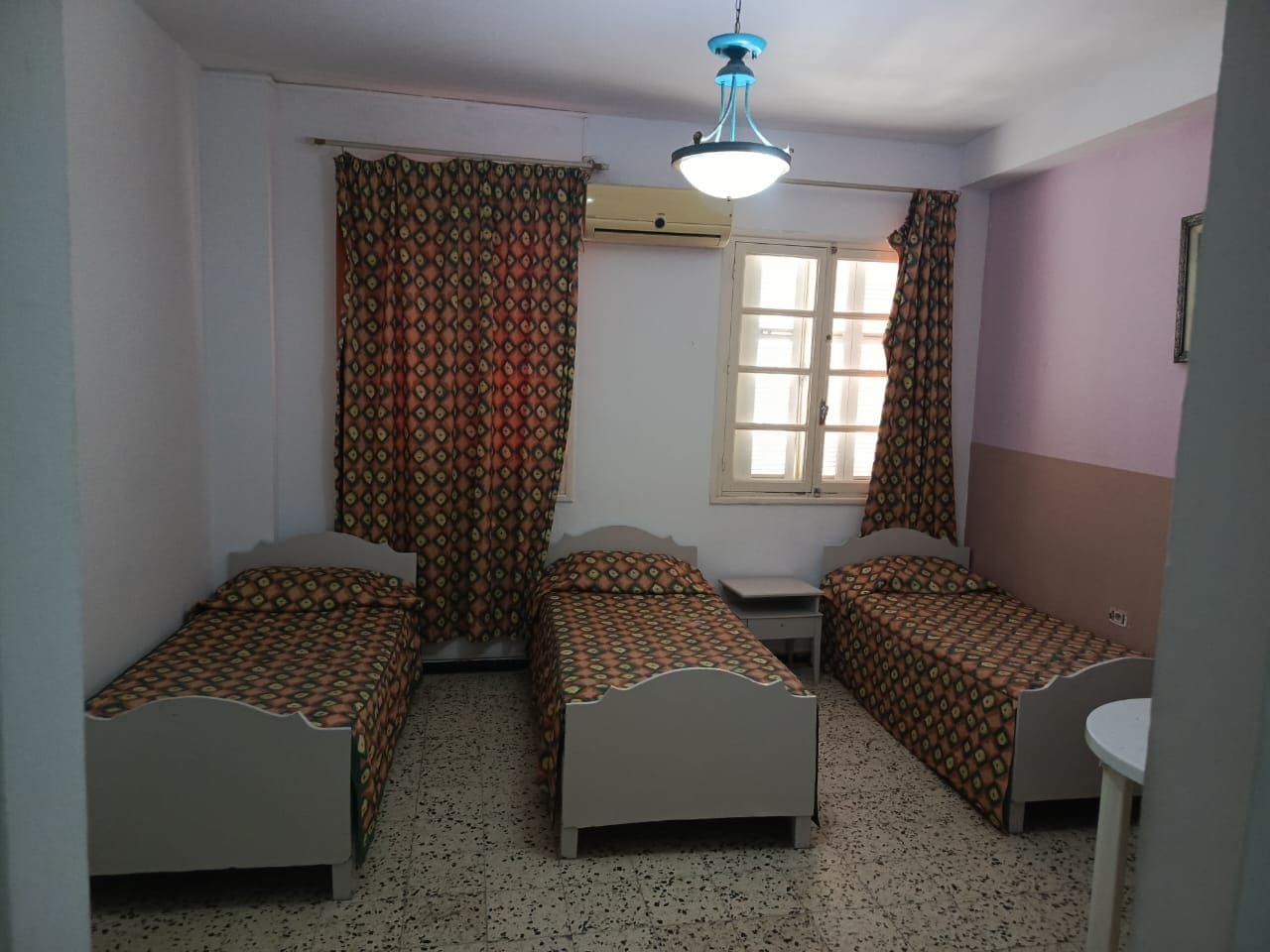 A room with three single beds, a window with curtains, a ceiling light, and a small table.