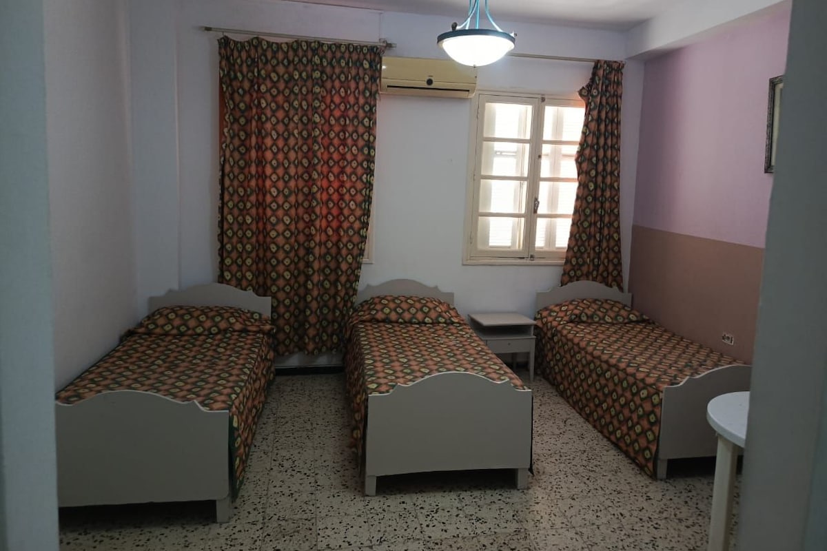 A room with three single beds, a window with curtains, a ceiling light, and a small table.