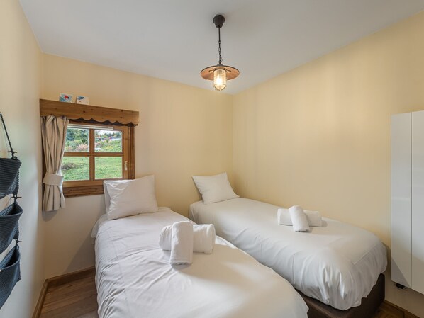 A room with two single beds, a wooden ceiling, a window with curtains, and a hanging light fixture.