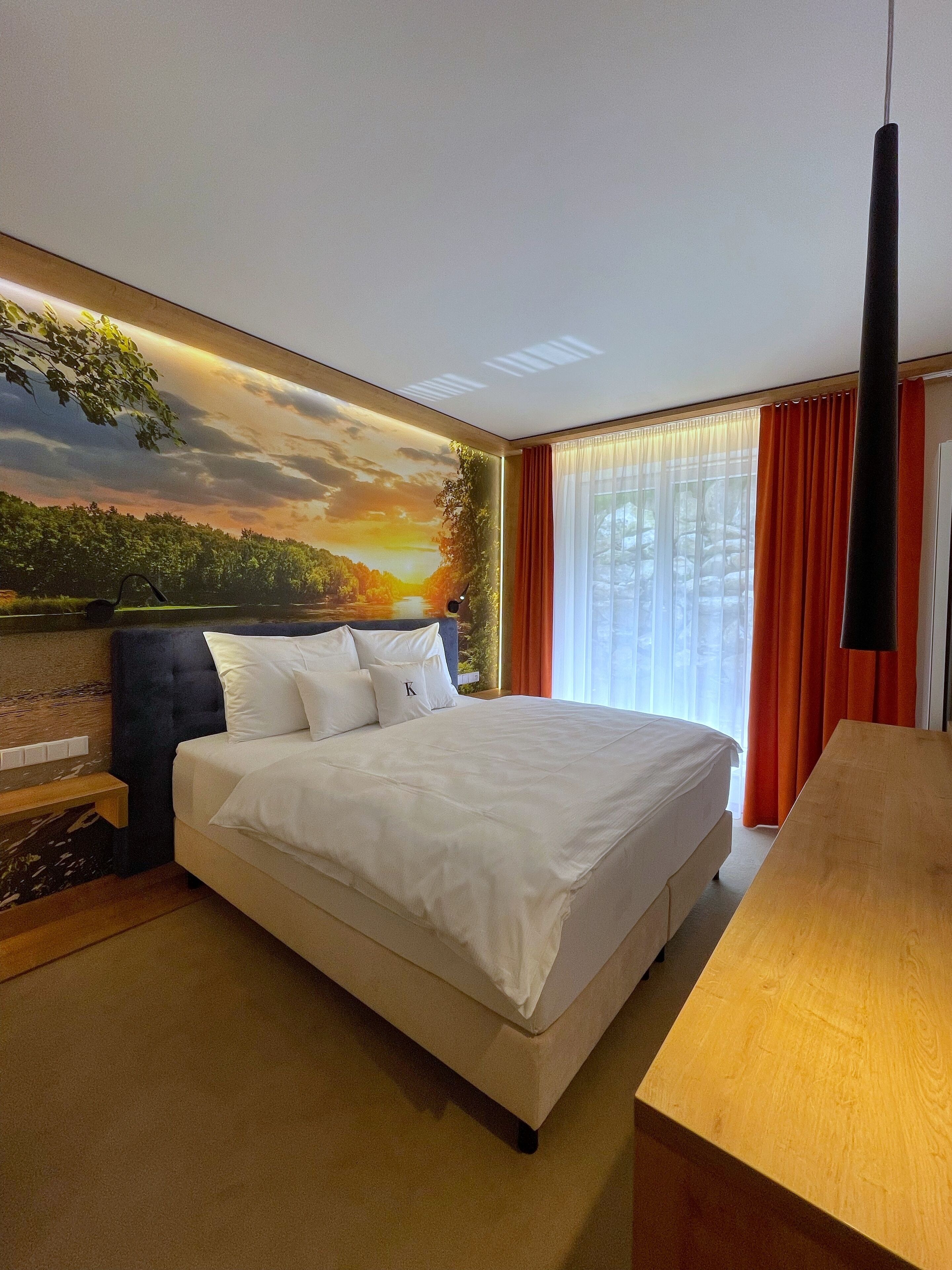 A hotel room with a large bed, a wooden nightstand, a painting of a sunset, and a view of the outdoors through the window.