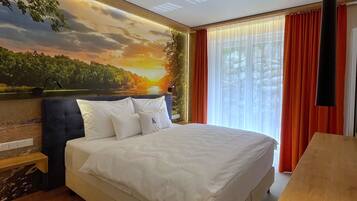 A hotel room with a large bed, a wooden nightstand, a painting of a sunset, and a view of the outdoors through the window.