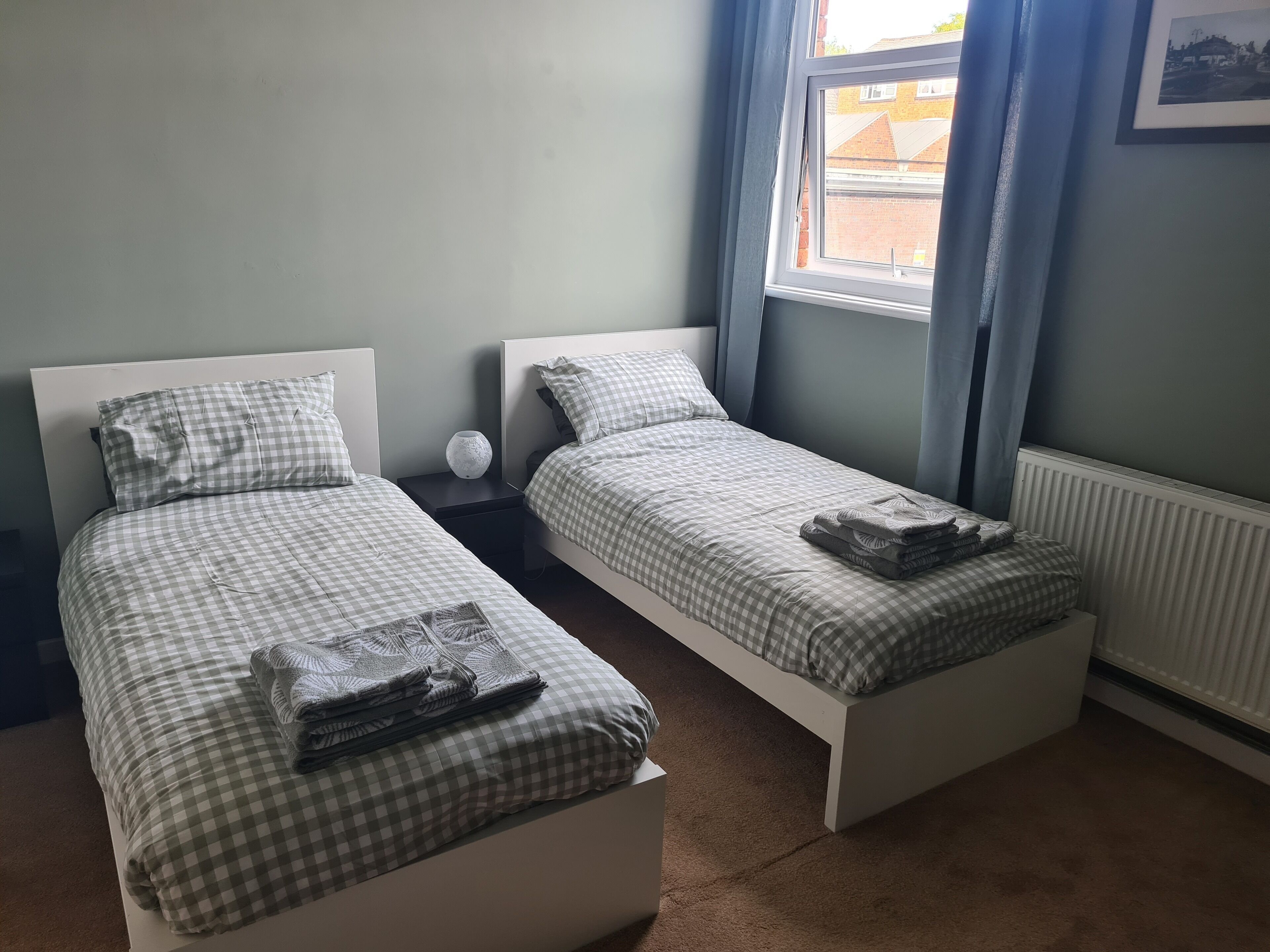 Un dormitorio doble con dos camas individuales, una mesita de noche, un jarrón y una ventana con cortinas.