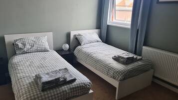 Un dormitorio doble con dos camas individuales, una mesita de noche, un jarrón y una ventana con cortinas.