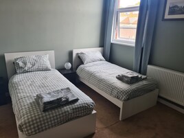 Un dormitorio doble con dos camas individuales, una mesita de noche, un jarrón y una ventana con cortinas.
