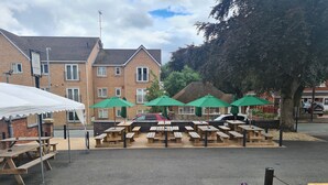 A picnic area with wooden tables and green umbrellas, a brick building in the background, and a car parked nearby. - The Hen House (Cannock)