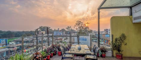 Una terraza en la azotea con mesas y sillas, plantas en macetas y vistas de la ciudad.
