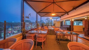 Un espacio exterior para comer con sillas y mesas de mimbre, una zona cubierta con vistas a la ciudad y una barra con taburetes.