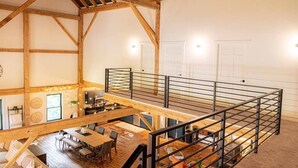 Ett rymligt rum i loftstil med högt i tak, synliga träbjälkar, en övre våningsplan och en matplats med ett långt bord och stolar.
