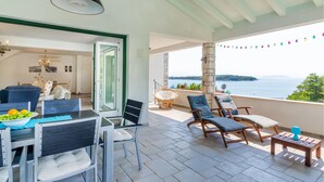 En balkon med udsigt over havet, møbleret med stole og et bord, samt et spiseområde med stole og en skål med frugt.