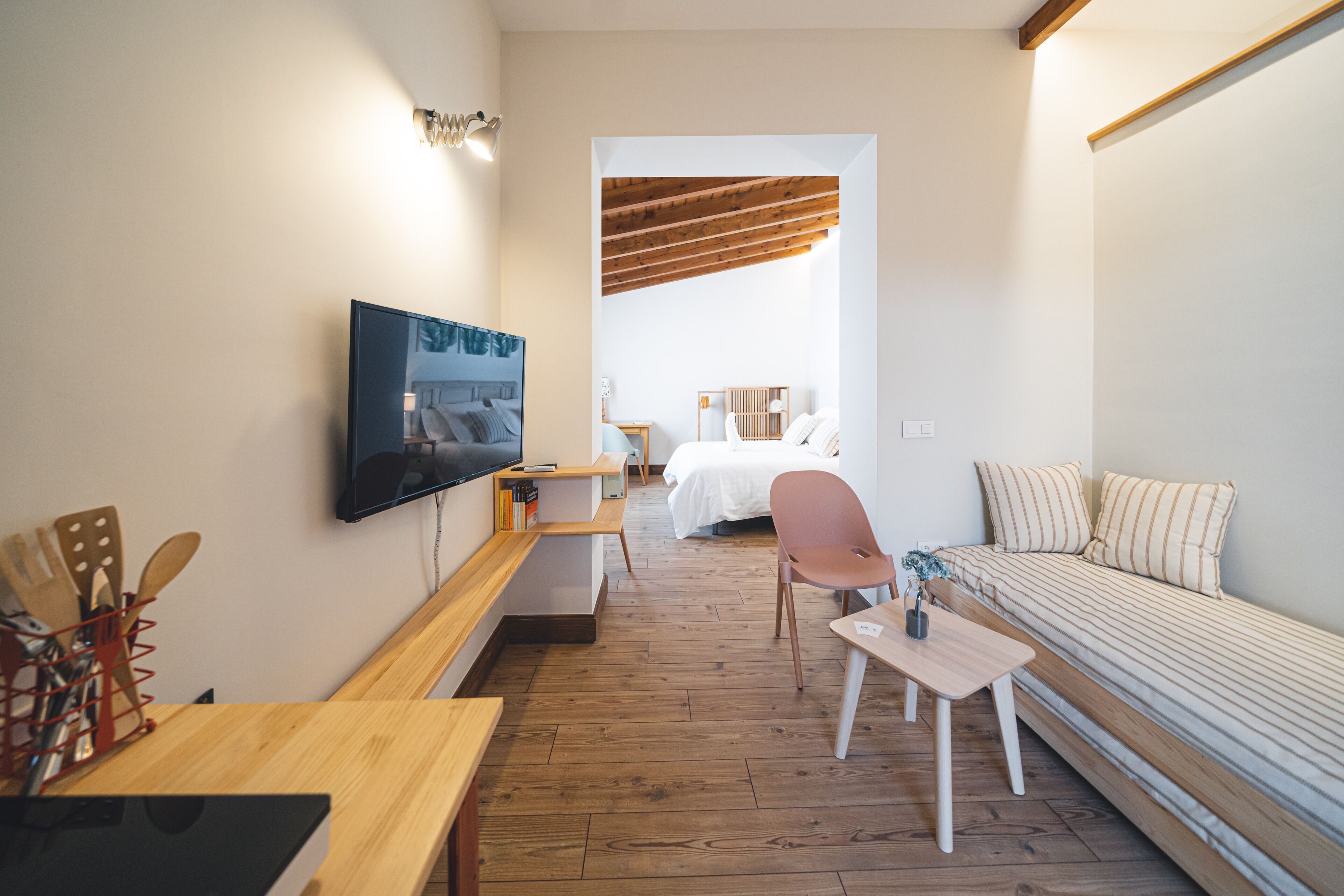A compact room with a bed, a TV mounted on the wall, a wooden table, a chair, and a small table with a vase.