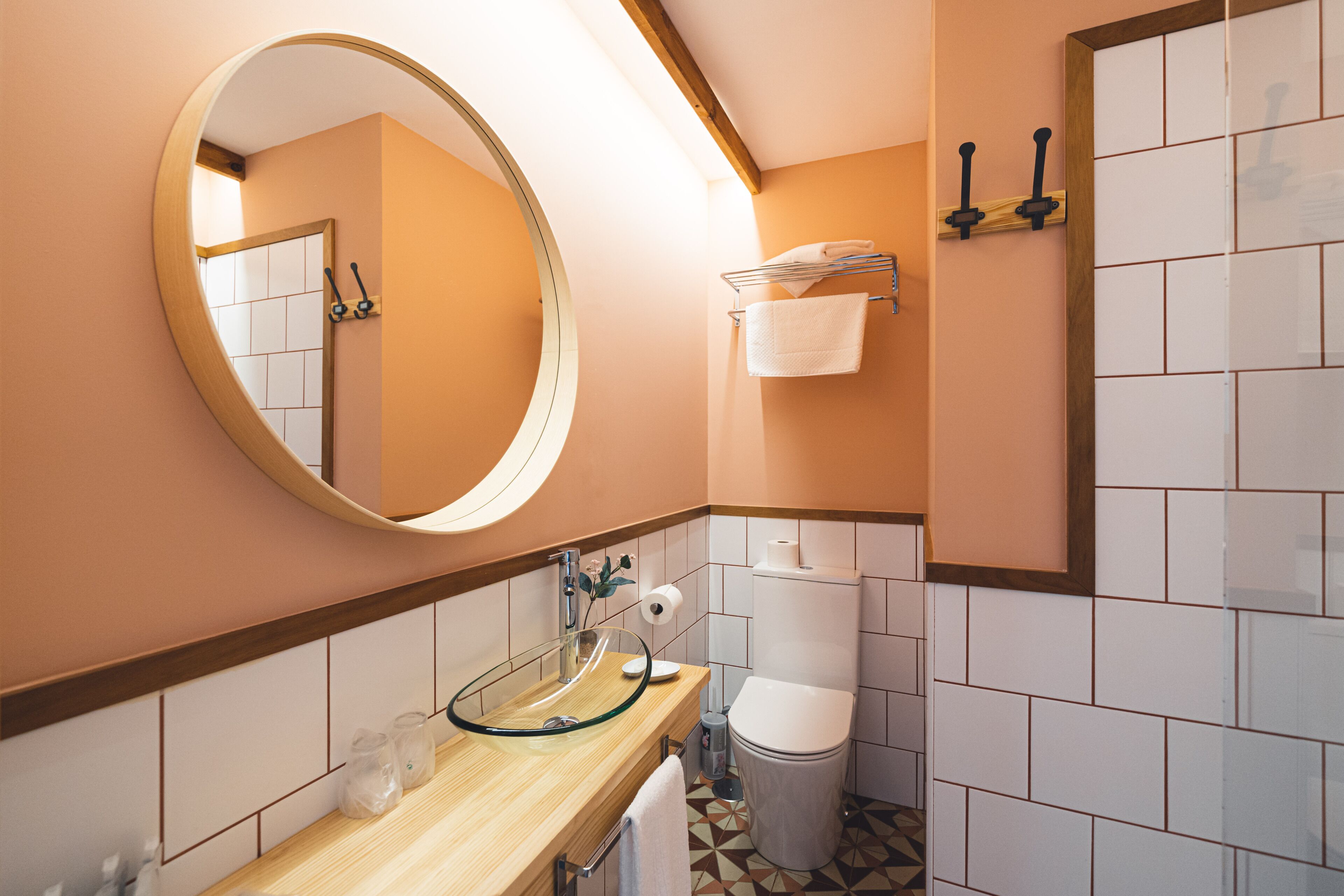 A modern bathroom with a round mirror, glass sink, white tiles, and a toilet.