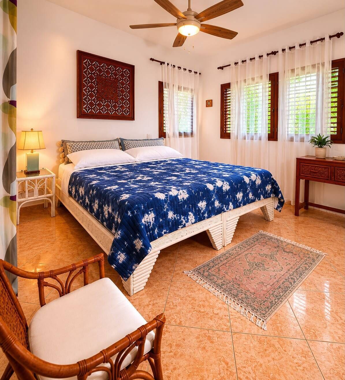 Une chambre à coucher comprenant un lit, une chaise, une table de chevet, un ventilateur de plafond et une fenêtre avec des rideaux.