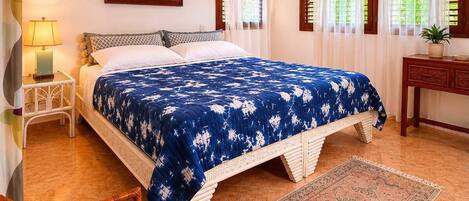 Une chambre à coucher comprenant un lit, une chaise, une table de chevet, un ventilateur de plafond et une fenêtre avec des rideaux.