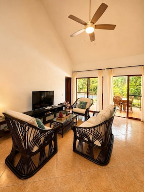 A spacious living room with a ceiling fan, a flat-screen TV, wicker furniture, and a view of greenery through large windows.