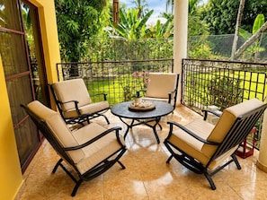 A patio with four chairs and a table, surrounded by a metal fence, with a view of a garden.