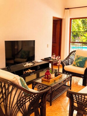 A living room with a television, wicker furniture, a glass coffee table, and a view of a pool.