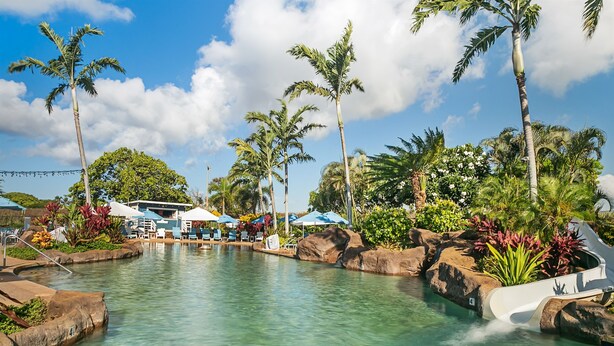 Uma área de piscina com espreguiçadeiras e guarda-sóis, rodeada por palmeiras e plantas tropicais.