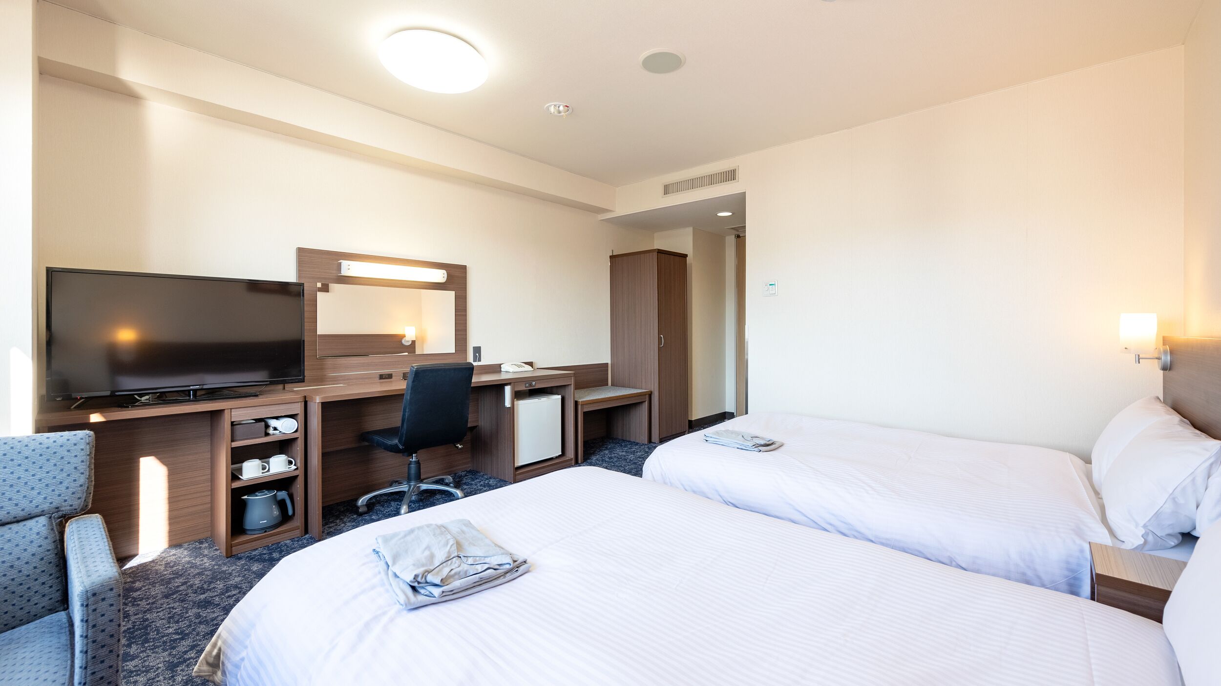 A hotel room with two beds, a desk with a computer, a television, and a minibar.