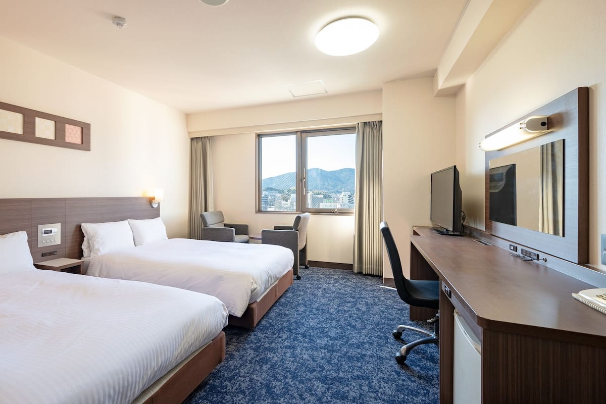 Habitación de hotel con dos camas, un escritorio con televisor, una silla y ventana con vista a la ciudad.
