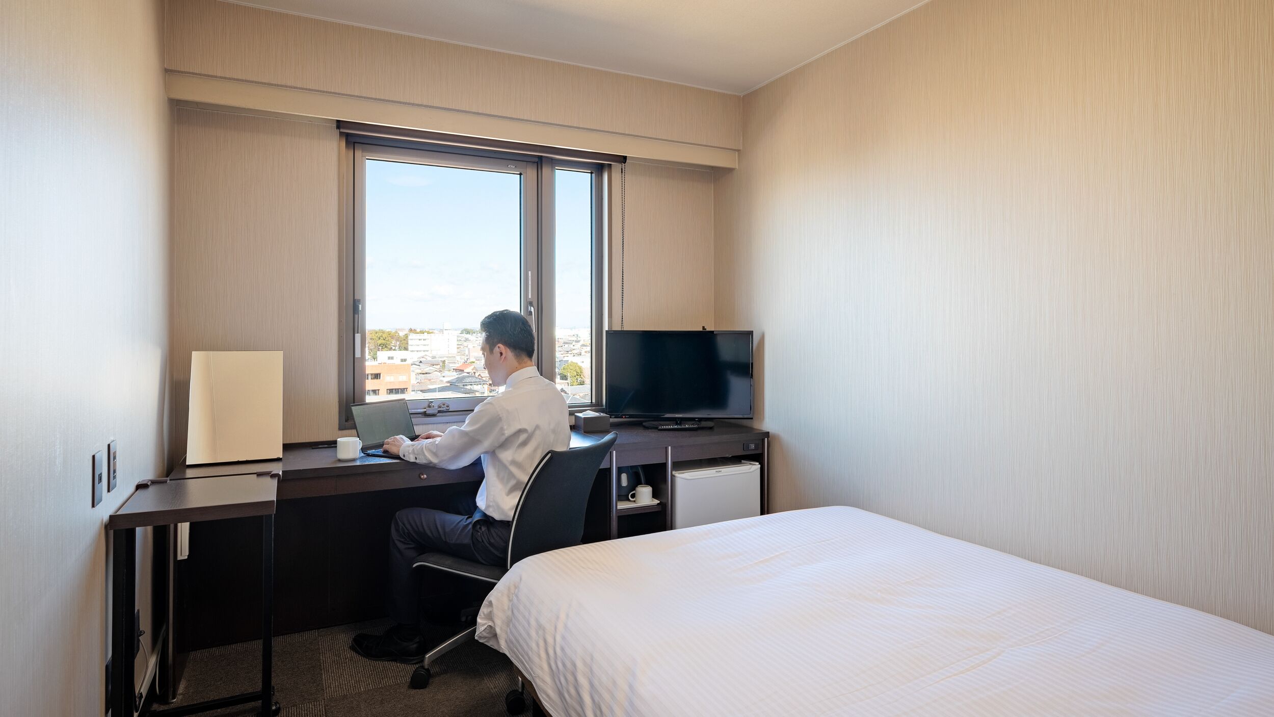 A hotel room with a bed, a desk with a computer, a TV, and a window with a city view.