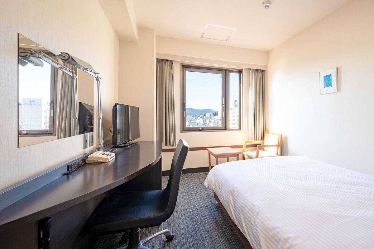 Habitación de hotel con cama, escritorio, ordenador, teléfono, silla y ventana con vista a edificios.