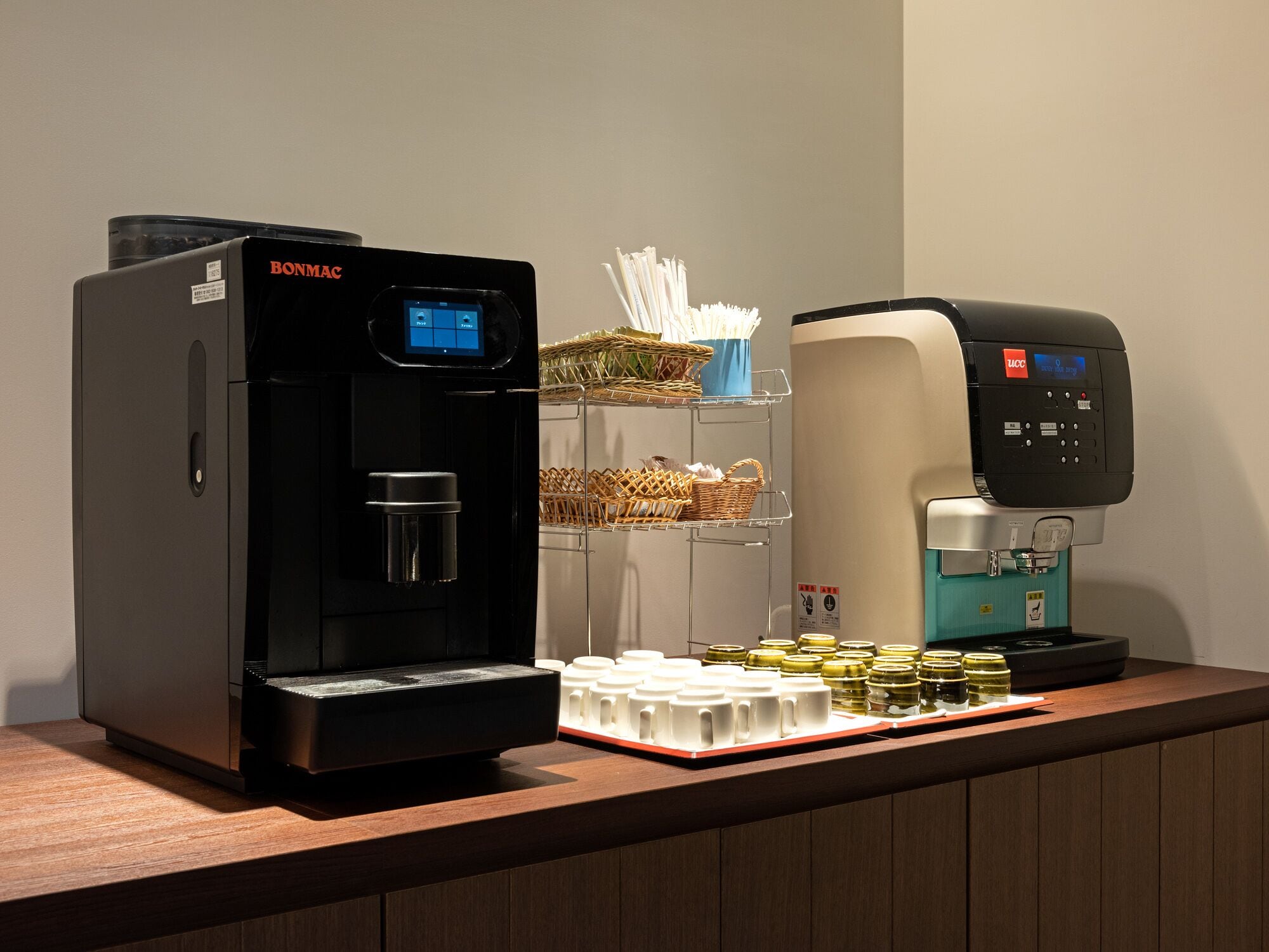 A coffee station with a black Bonmac coffee machine, a blue LCC coffee machine, and a tray of disposable cups and condiments.