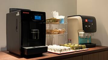 A coffee station with a black Bonmac coffee machine, a blue LCC coffee machine, and a tray of disposable cups and condiments.