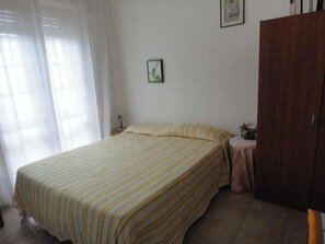 Un dormitorio con cama, mesita de noche, lámpara, armario y ventana con cortinas.