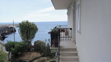 Eine Treppe führt zu einem Balkon mit Blick auf eine zerklüftete Küstenlinie, eine Kaktusstaude und Kübelpflanzen.