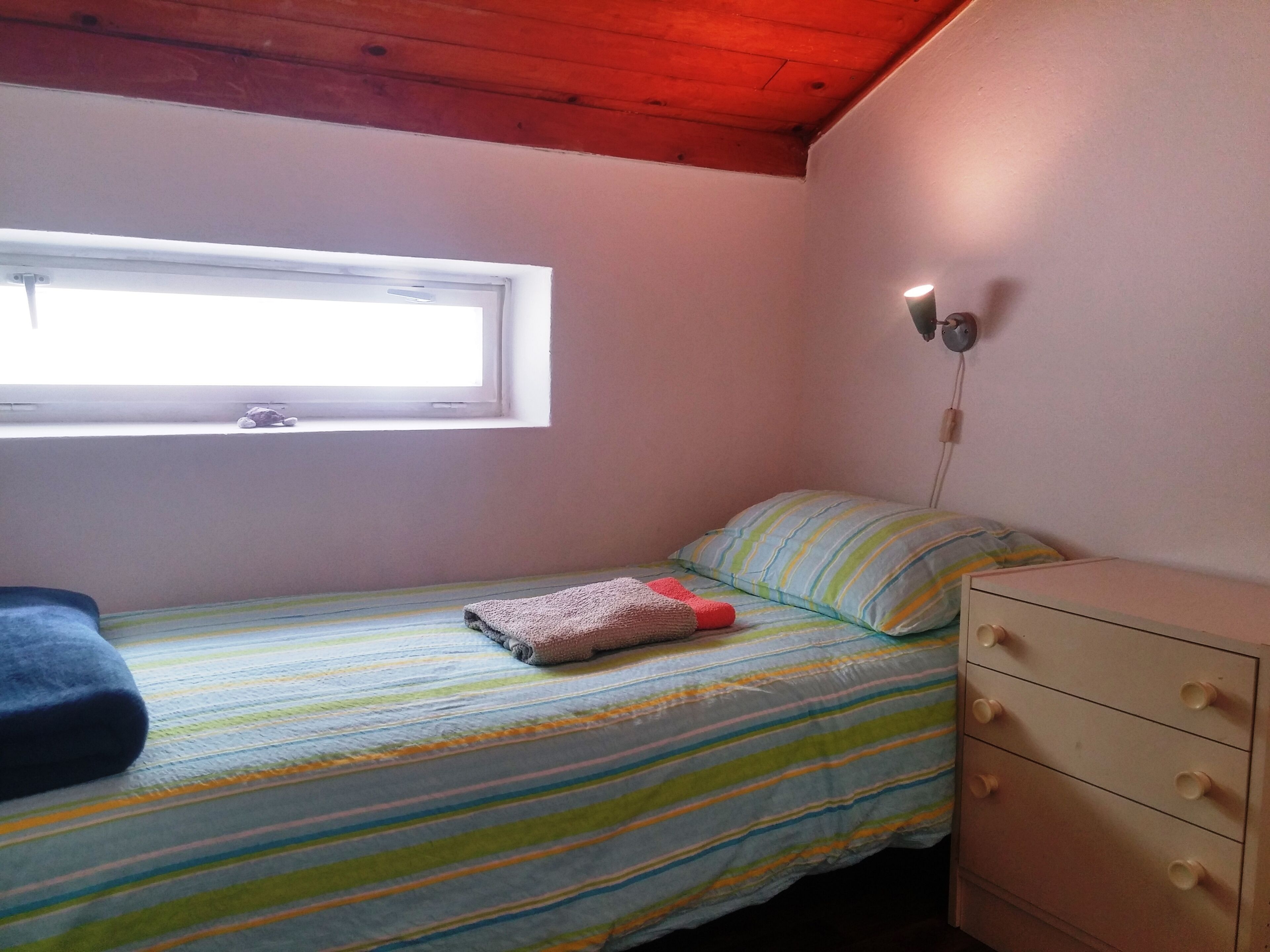 Une petite chambre avec un lit simple, une fenêtre, une lampe fixée au mur et une commode.