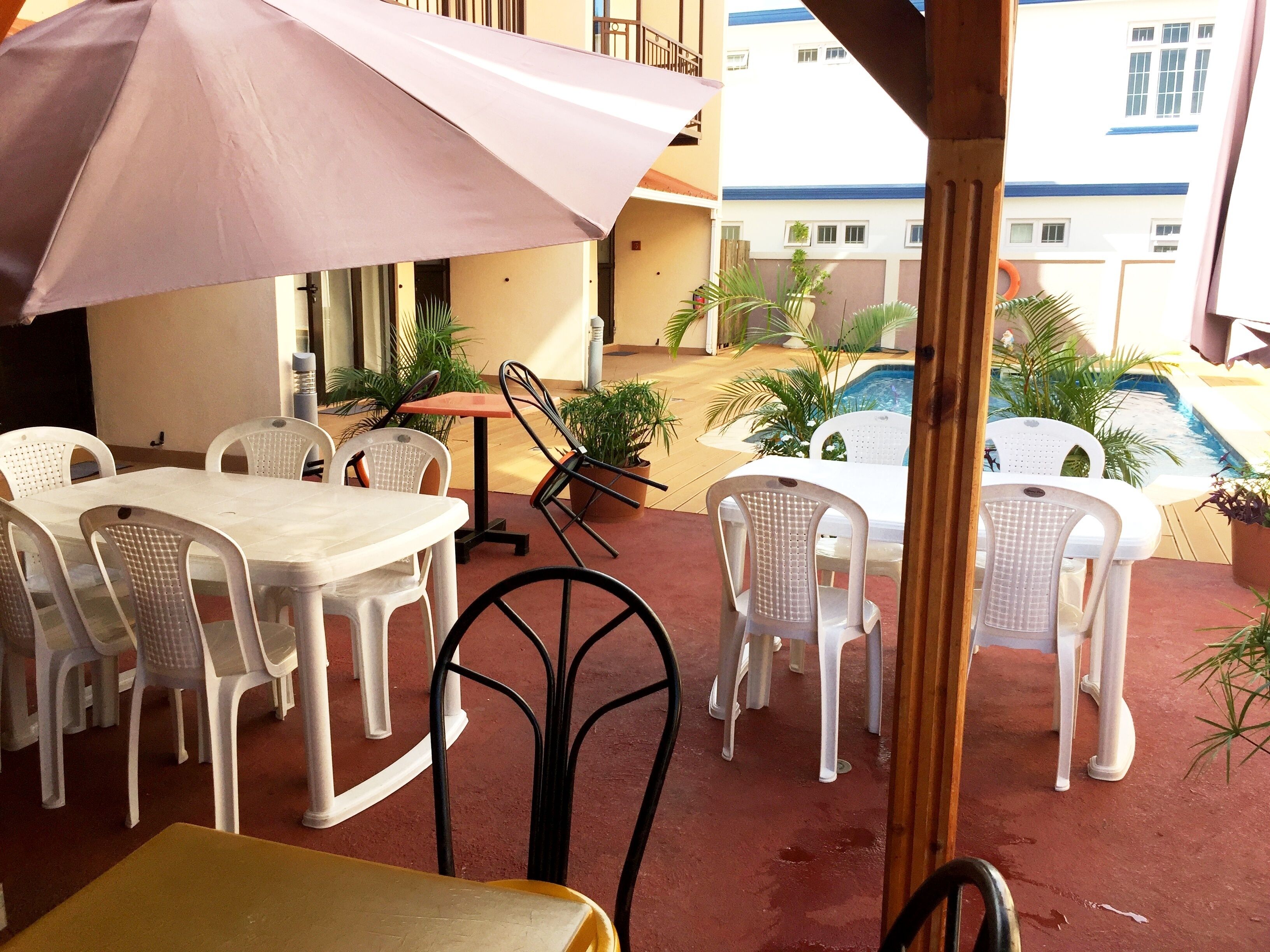 Une terrasse extérieure avec des chaises et des tables en plastique blanc, un parasol rose et une vue sur une piscine et des bâtiments en arrière-plan.