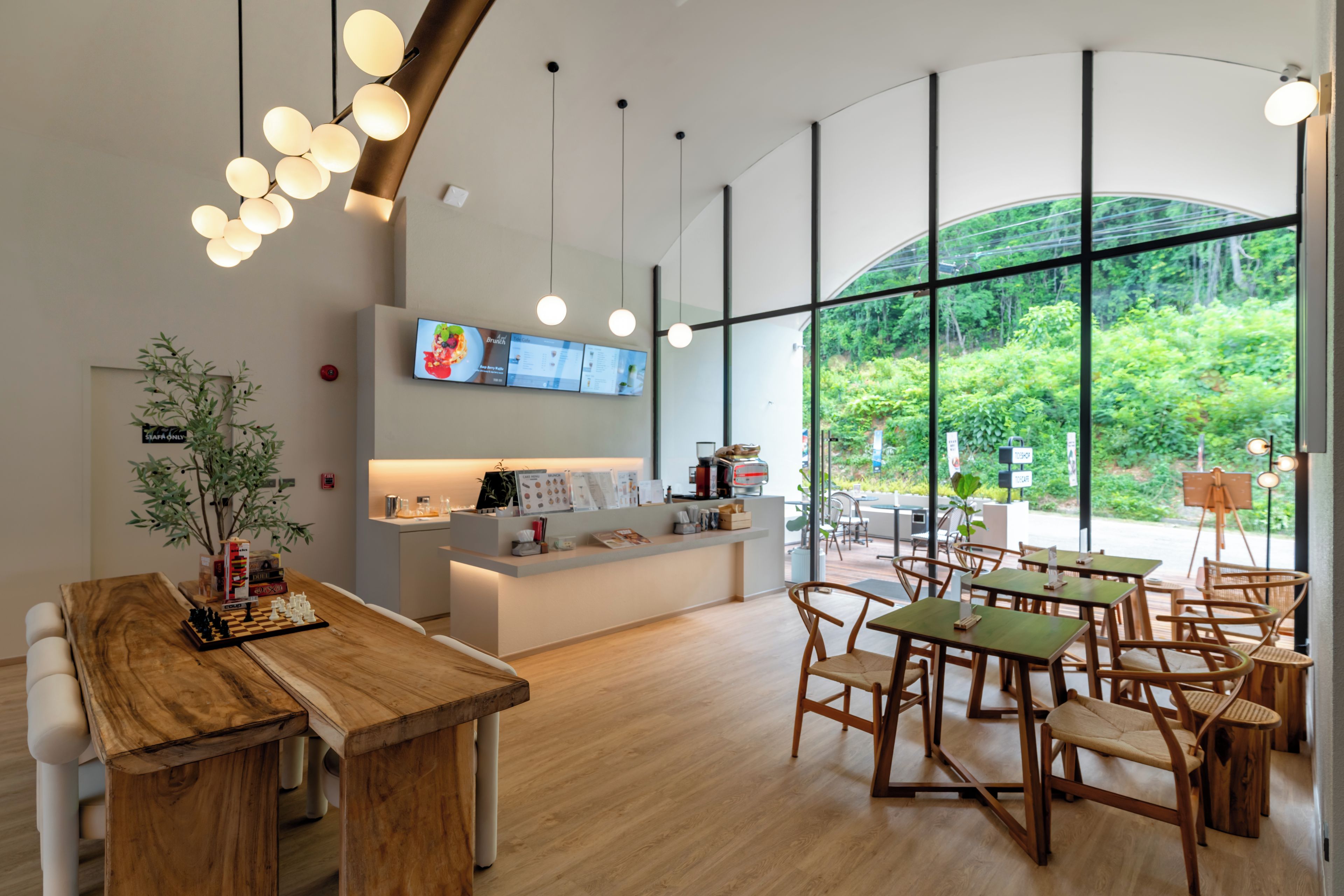 Een modern café interieur met houten tafels en stoelen, een balie met een menukaart en grote ramen met uitzicht op groen.