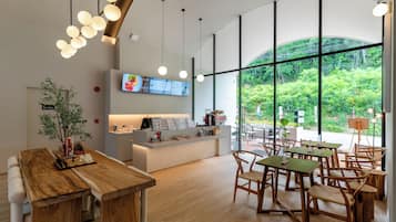 A modern cafe interior with wooden tables and chairs, a counter with a menu, and large windows offering a view of greenery.