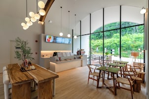 Interior de una cafetería moderna con mesas y sillas de madera, una barra con un menú y amplios ventanales con vistas a zonas verdes.