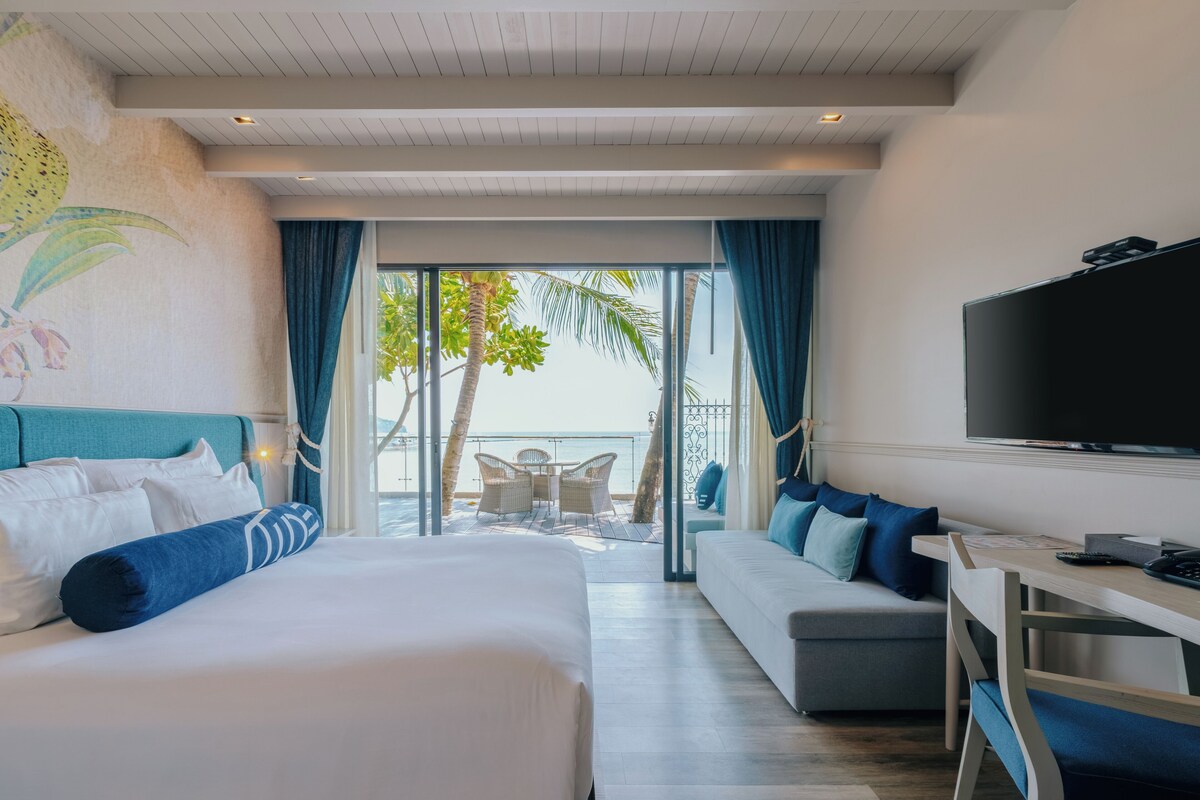 Een hotelkamer met een bed, een bank, een televisie en een balkon met uitzicht op palmbomen.