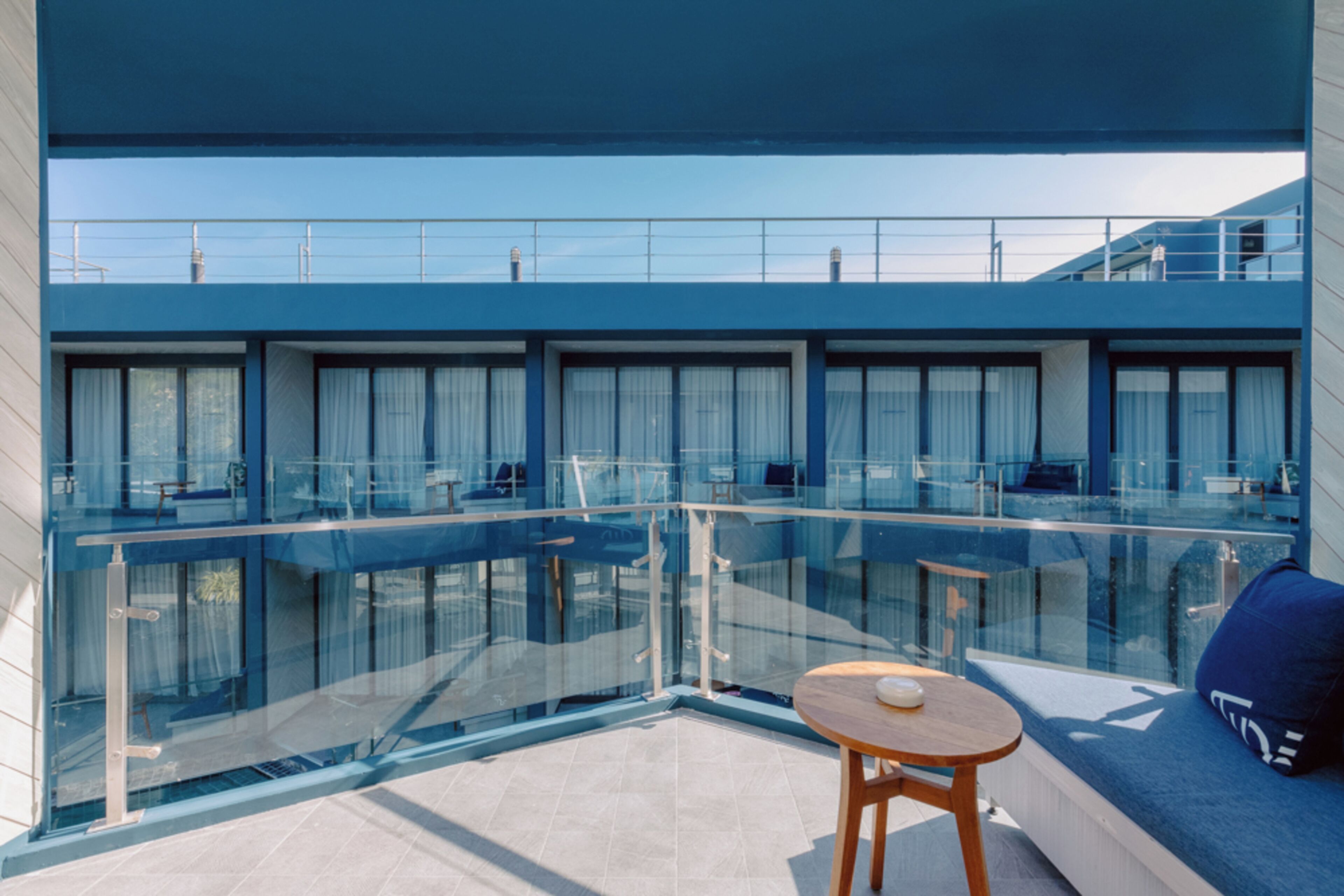 Un balcon moderne doté d’une balustrade en verre, d’une table ronde en bois et d’un coin salon confortable agrémenté d’un coussin bleu.