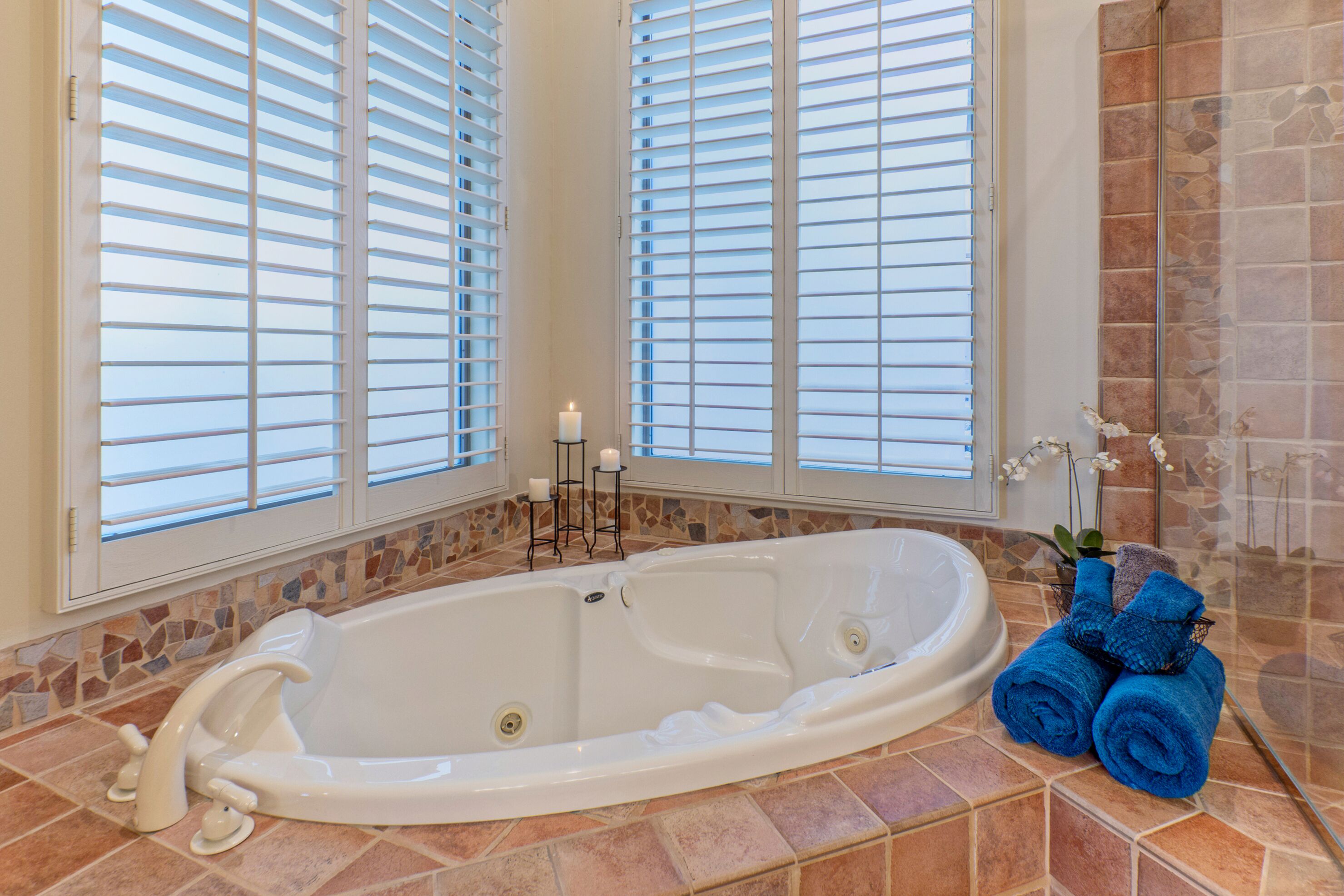 A bathroom with a large bathtub, tiled floor, and windows with blinds.