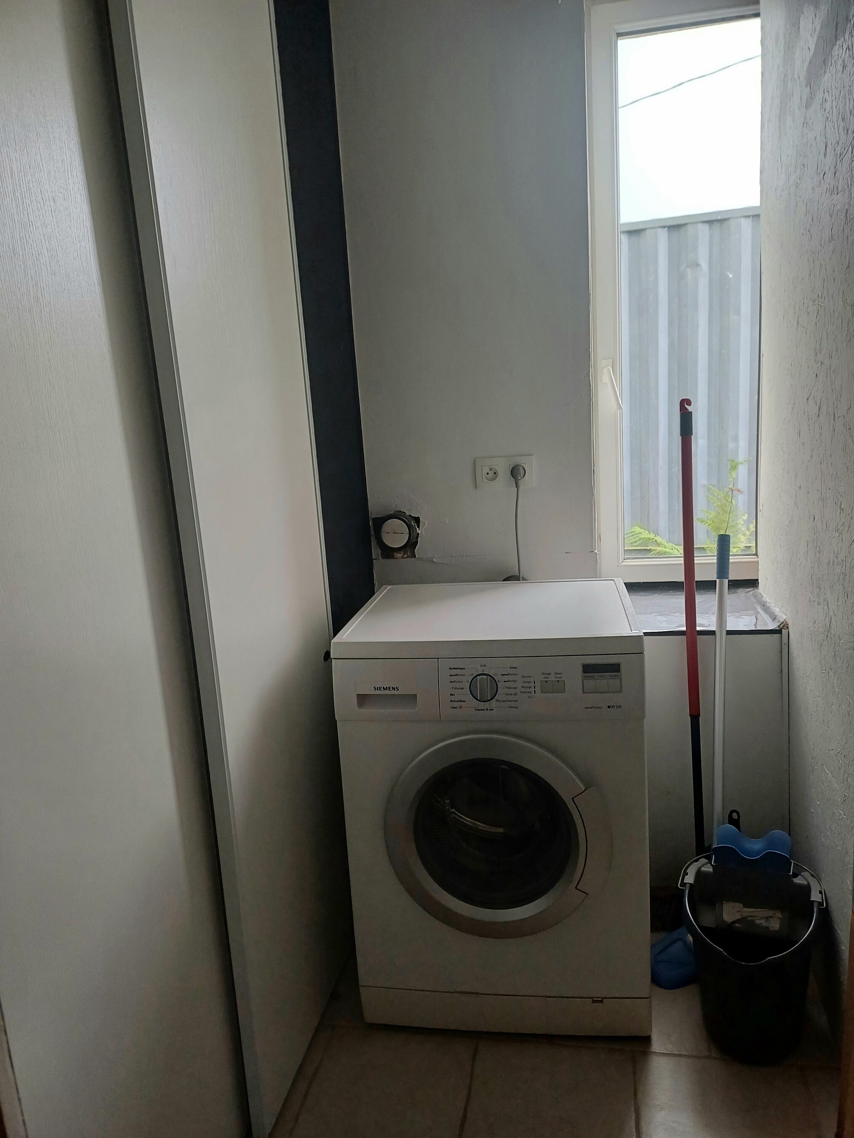Ein kleiner Waschraum mit einer Frontlader-Waschmaschine, einer Steckdose, einem Fenster mit Ausblick und einem an die Wand gelehnten Mop.